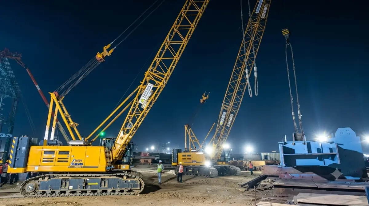 Two crawler cranes rigged for a night lift under floodlights, with erection crew standing by